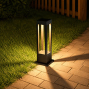 Black rectangular garden path light glowing on grass beside a brick walkway.