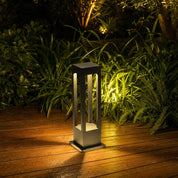 Black rectangular outdoor bollard light glowing warmly on a wooden deck beside green garden foliage.