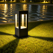 Black rectangular outdoor bollard light glowing on grass beside rippling water.