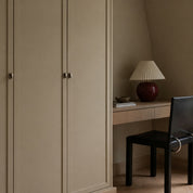 Beige wardrobe with small knobs beside a wooden desk with lamp and black chair.