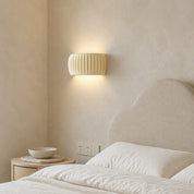 Cream ribbed wall sconce glowing above a neutral upholstered bed in a cozy bedroom.