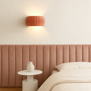 Terracotta ribbed wall sconce glowing above a pink upholstered bed headboard.