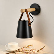 Black wall lamp with wood arm and brown leather straps above a white cup and blossoms.