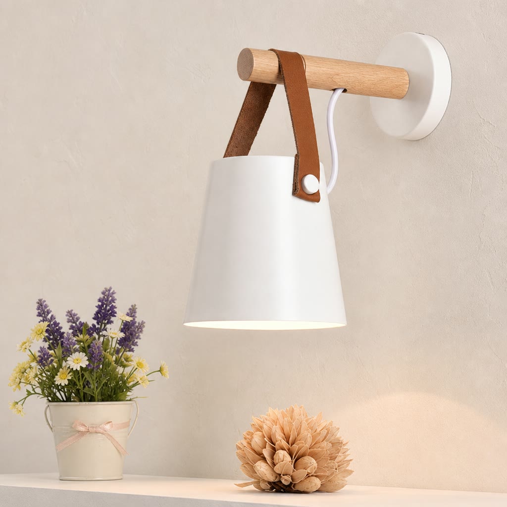 White wall lamp with wooden peg and brown leather straps above small flowers.