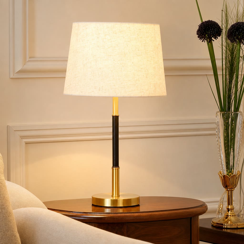 Lit table lamp with a cream fabric shade, black and brass stem, and round brass base on a wooden table.