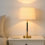 Lit table lamp with beige shade, black and gold stem, and round gold base on a white nightstand.