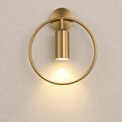 Brushed gold circular wall sconce with a glowing downward cylinder light on a pale wall.