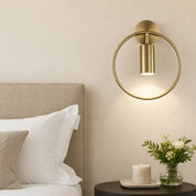 Gold circular wall sconce with a cylindrical downlight above a bedside table.