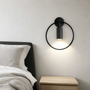 Black circular wall sconce with a downward cylinder light above a wooden bedside table.