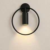 Black circular wall sconce with a cylindrical downlight casting a warm glow on a pale wall.