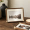 Wood and brass photo frame with white mat displaying a mountain landscape on a rustic table.