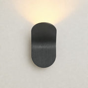 Black rounded wall sconce casting a warm upward glow on a pale wall.
