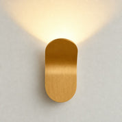 Gold capsule wall sconce with warm upward glow on a pale wall.