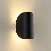 Black half-cylinder wall sconce casting warm light on a textured neutral wall.