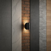 Black cylindrical wall sconce glowing on a brick panel between vertical material samples.