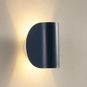 Dark half-cylinder wall sconce glowing warmly on a neutral plaster wall.
