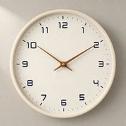 Minimal white wall clock with black numerals and wooden hands on a pale wall.