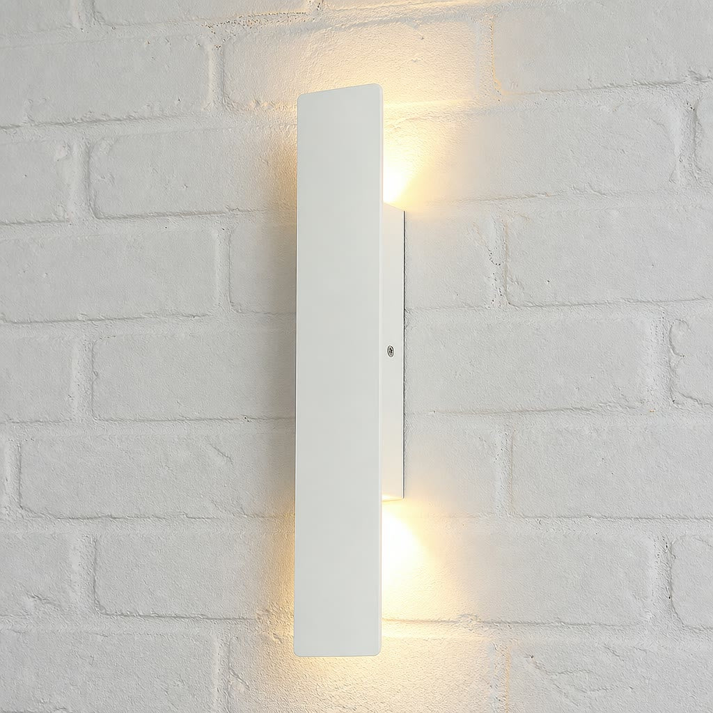 Slim white vertical wall light glowing warmly on a white brick wall.