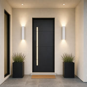 Black modern front door with vertical light panel, flanked by wall sconces and square planters.