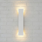 Slim white rectangular wall sconce glowing warmly on a pale textured brick wall.