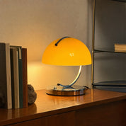Yellow dome table lamp with chrome base glowing on a wooden shelf.