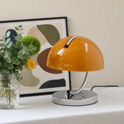 Glossy orange dome table lamp with chrome base on a white table beside flowers and framed art.