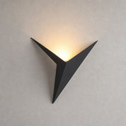 Black triangular wall sconce casting a warm upward glow on a pale textured wall.