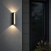 Rectangular black outdoor wall light glowing on a textured wall beside a dark front door.