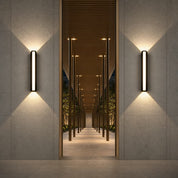 Two black vertical wall sconces cast warm light on textured walls flanking a modern corridor.
