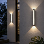 Slim black vertical wall sconce casting warm up-and-down light on a textured exterior wall.