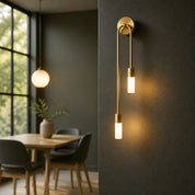 Brass double pendant wall sconce with two glowing cylindrical bulbs on a dark textured wall.