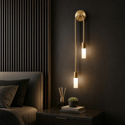 Brass dual-cylinder wall sconce glowing beside a bed and nightstand.
