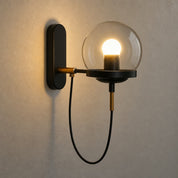 Modern wall sconce with a glowing globe shade, black mount, brass accents, and looped black cord.
