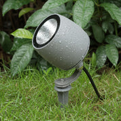 Gray outdoor spotlight with water droplets mounted on a stake in wet grass.