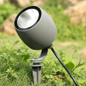 Gray outdoor spotlight on a ground stake among green grass and small plants.