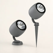 Two gray cylindrical outdoor spotlights with black rims and silver reflectors, one on a base and one on a ground spike.