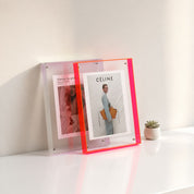 Neon pink acrylic photo frame holding fashion prints on a glossy white shelf.