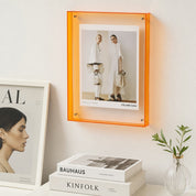 Orange acrylic wall frame displaying a Celine fashion poster above white books and a small plant.
