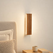 Wooden wall sconce with a warm vertical light beside a bed and nightstand.