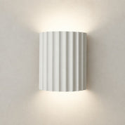 White ribbed cylindrical wall sconce glowing warmly on a plain wall