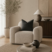 Cream boucle lounge chair with rounded arms and a dark charcoal pillow in a warm neutral living room.