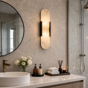 Warm vertical frosted wall sconce with dark side brackets above a marble bathroom vanity.