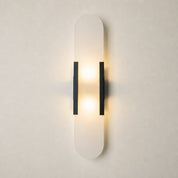 Vertical frosted wall sconce with black side brackets glowing warmly on a pale wall.