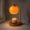 A brass candle warmer lamp with an orange glass shade illuminates a jar candle on a wooden base.