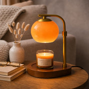 Elegant glass candle warmer lamp casting a soft orange glow onto a lit candle on a wooden table, next to books and a vase.
