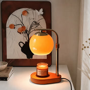 An elegant glass candle warmer lamp casting a soft orange glow, placed on a wooden base with a lit sandalwood and smoke candle.