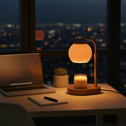 An elegant glass candle warmer lamp emitting a soft orange glow on a desk next to a laptop and a small potted plant.