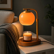 Elegant glass candle warmer lamp with a soft orange glow melting a jar candle on a white table next to a sofa.
