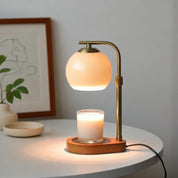 A brass candle warmer lamp with an orange glass shade sits on a table, warming a white candle on its wooden base.
