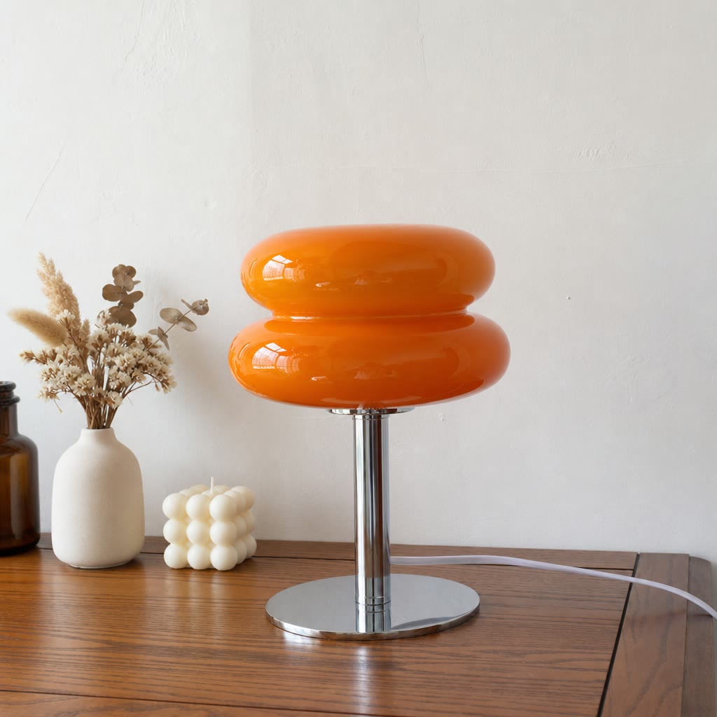 Glossy orange double-donut table lamp with chrome base on a wooden surface against a pale wall.
